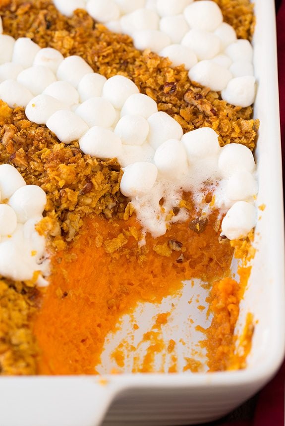 Browned Butter Sweet Potato Casserole Sweet Potato in a white casserole dish. One scoop is removed to show sweet potato layer. It's topped with rows of crushed cornflakes and pecans then alternating rows of mini marshmallows.