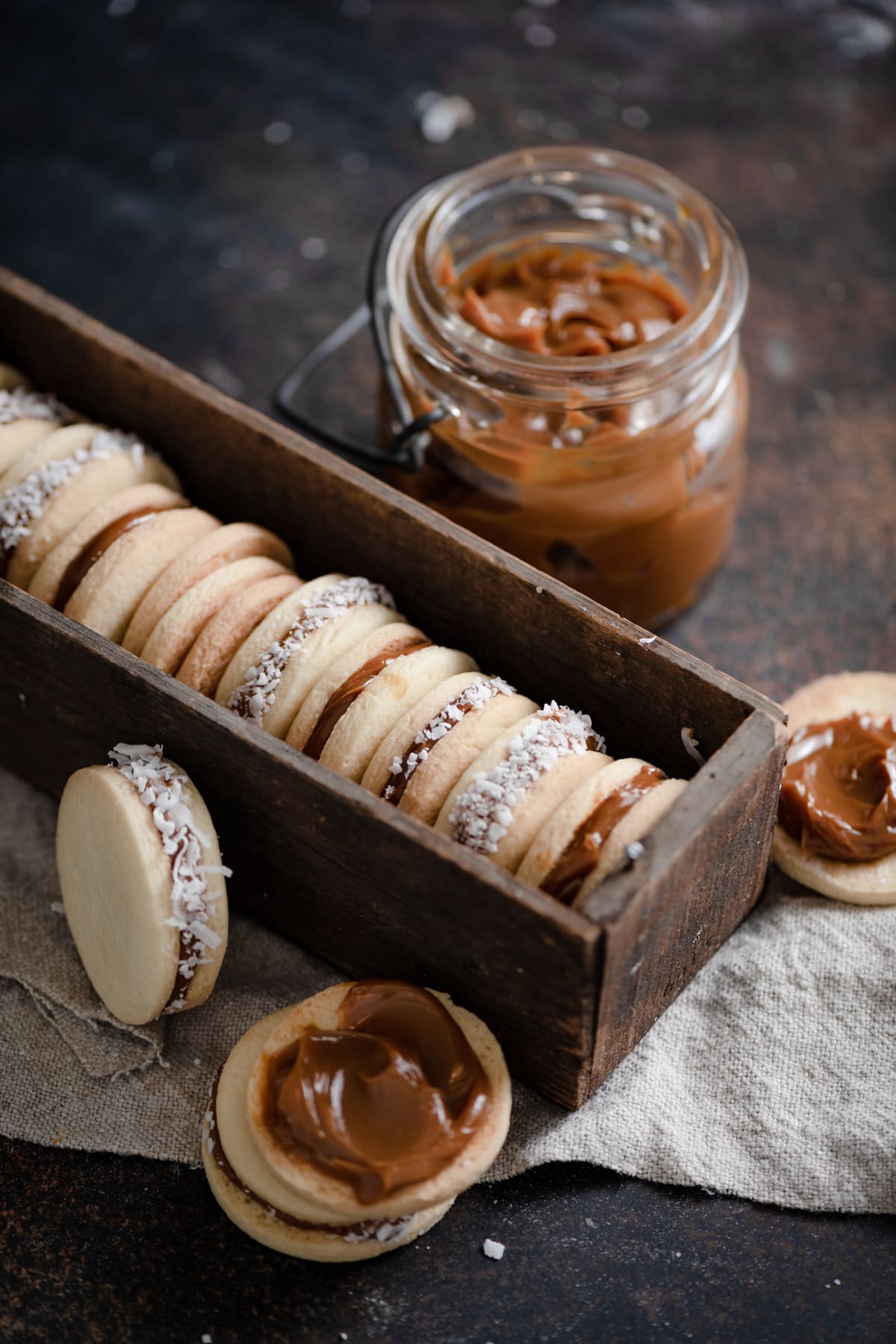 Alfajores Cookies {Dulce de Leche Sandwich Cookies} Alfajores Cookies in a box set on a cloth with dulce de leche in the background.