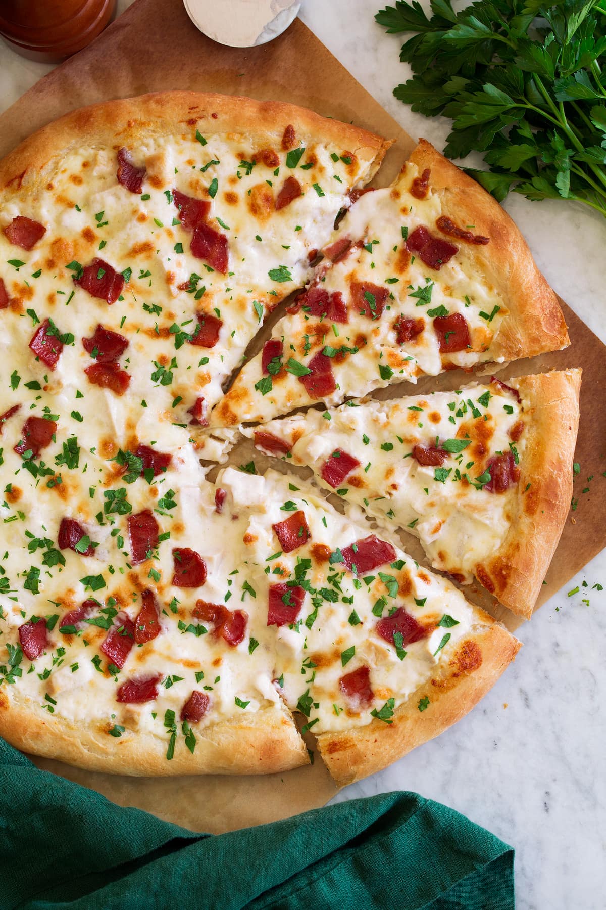 Chicken Alfredo Pizza Overhead photo of chicken alfredo pizza cut into slices. Pizza is set over parchment paper and has a green cloth to the side.