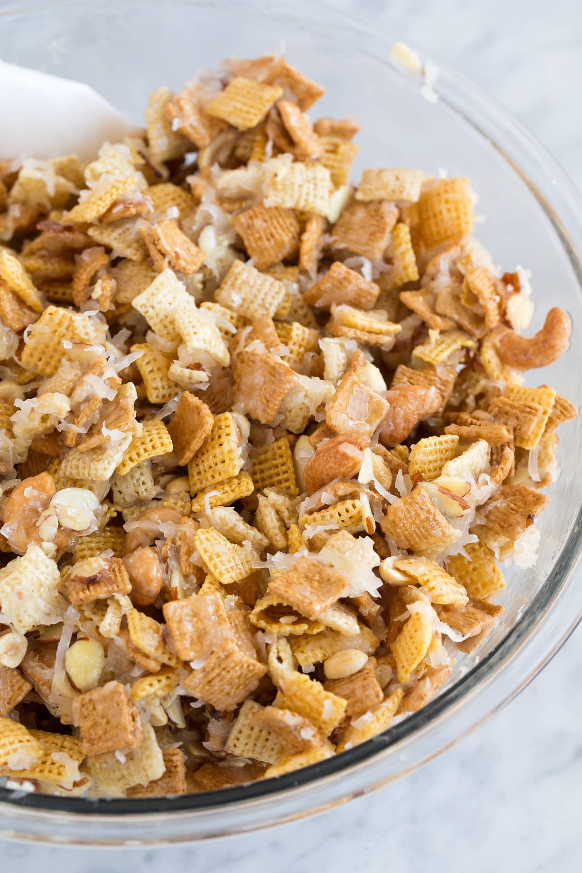 Sweet Holiday Chex Mix Sweet and sticky chex mix in a glass mixing bowl set over a marble surface. It includes corn and rice chex cereal, golden grahams, nuts, coconut and a butter sugar topping.