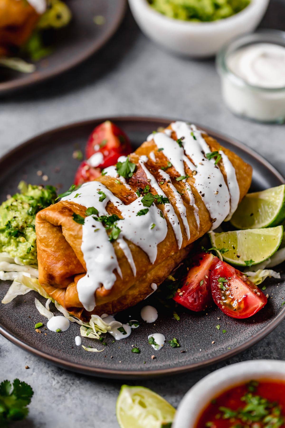 Chicken Chimichangas - one of the ultimate comfort foods! Perfectly hearty and layered with delicious ingredients like seasoned chicken, rich refried beans and shredded Mexican cheese. They're perfectly crisp and oh so satisfying! #chimichanga #chicken #mexican #recipe #refriedbeans #dinnerideas Homemade Chicken Chimichanga on a plate. It's drizzled with sour cream, garnished with cilantro and served with tomatoes and guacamole on the side.