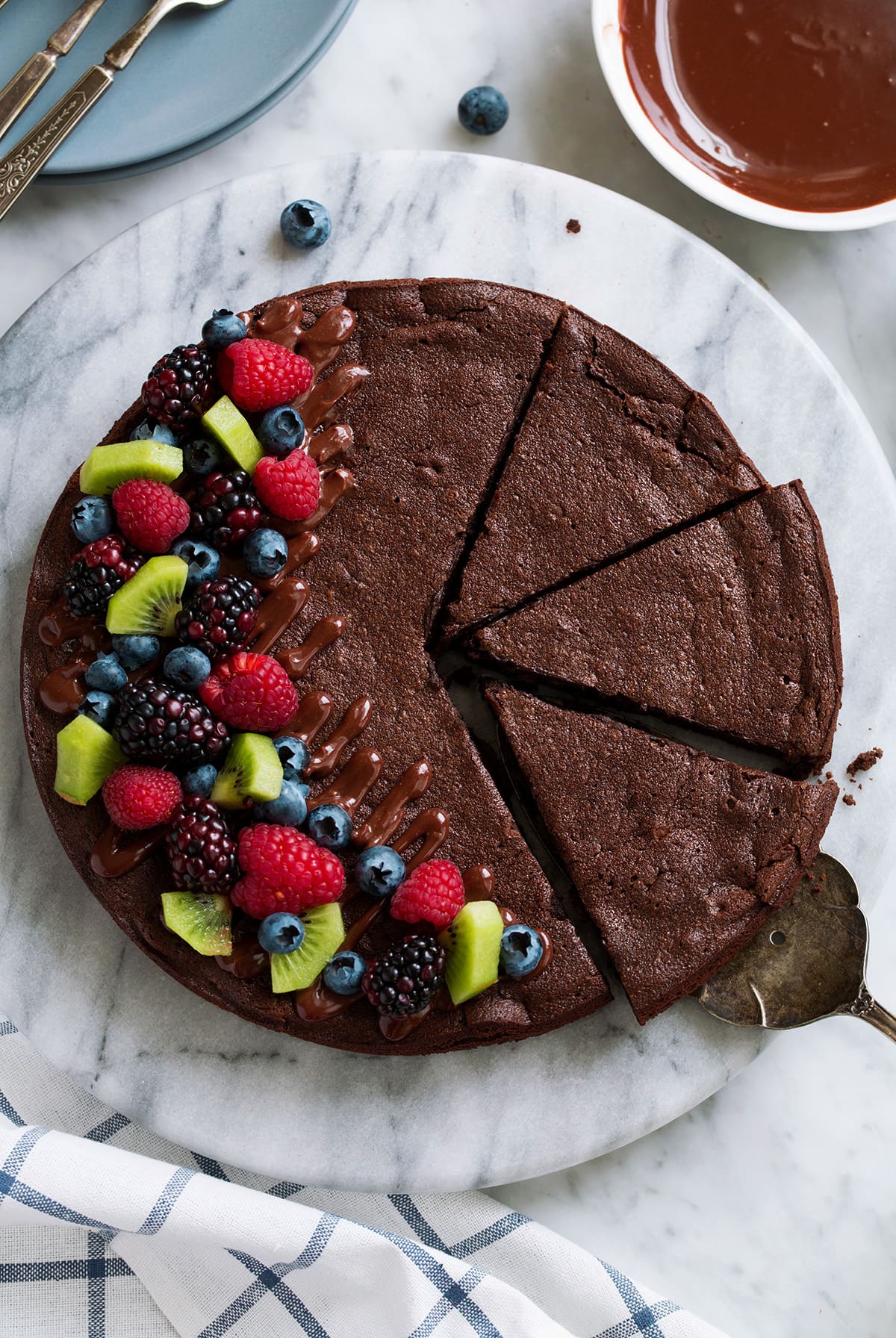 The BEST Flourless Chocolate Cake! Overhead image of flourless chocolate cake on a marble platter. Cake is topped with chocolate ganache drizzle and fresh fruit on the side.
