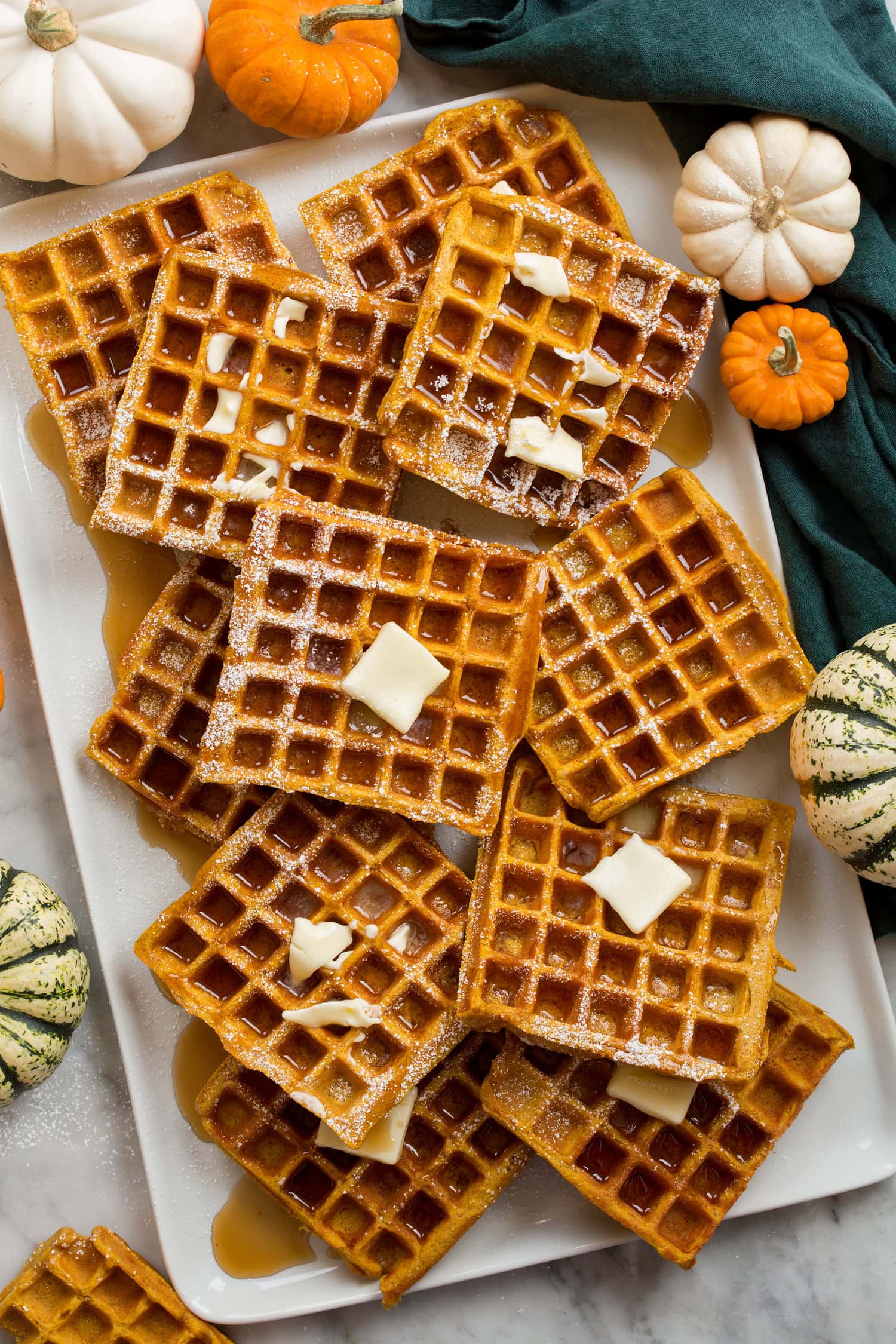 Pumpkin Waffles Pumpkin waffles layered on a white rectangular platter covered with syrup and butter, shown from above with pumpkins to the side.