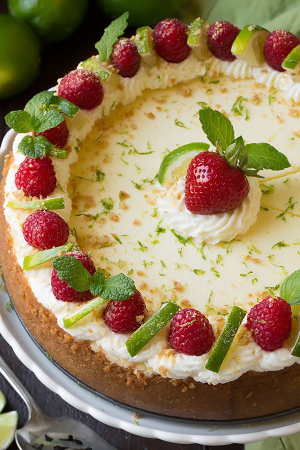 Key Lime Cheesecake Whole Key Lime Cheesecake on a white serving plate. Cheesecake is garnished with whipped cream, raspberries, and limes along edges and a strawberry in center.