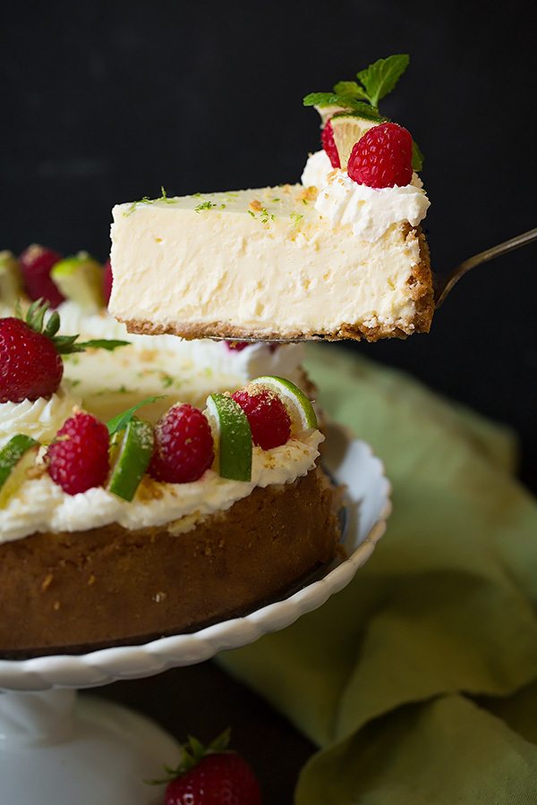 Key Lime Cheesecake Lifting a slice of key lime cheesecake from the whole cake. It's garnished with whipped cream, raspberries, mint and graham crackers.