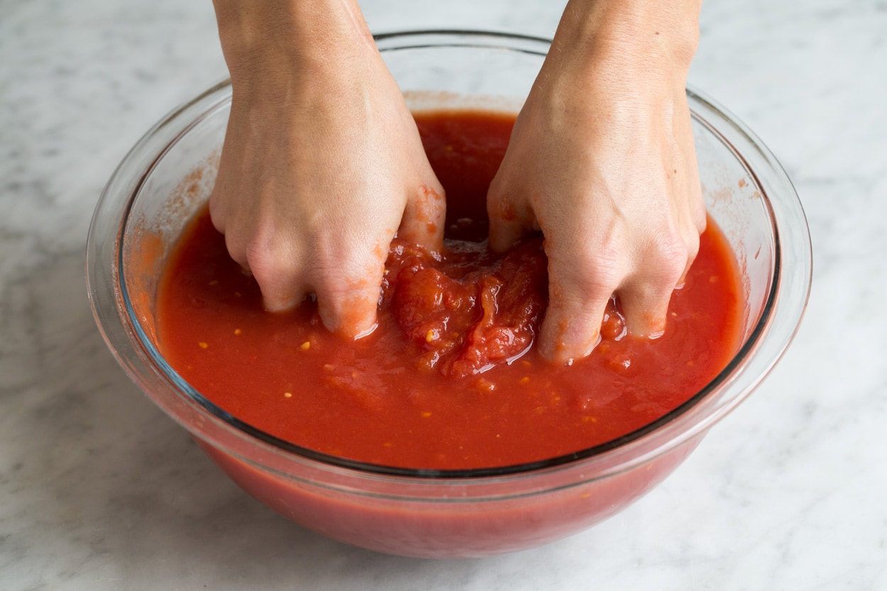 Homemade Marinara Sauce - so much better than the bottled stuff! This is authentic, fresh tasting sauce that you'll want to make again and again! Perfect over just about any type of pasta or served with cheese tortellini. It freezes really well too! #marinara #sauce #recipe #italianfood #dinner #lunch Crushing tomatoes by hand in a glass mixing bowl. Showing how to make marinara sauce.