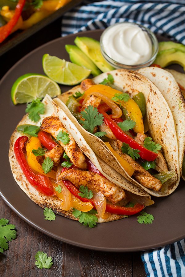 Chicken Fajitas Chicken Fajitas wrapped in tortillas served with limes and sour cream on brown plate.