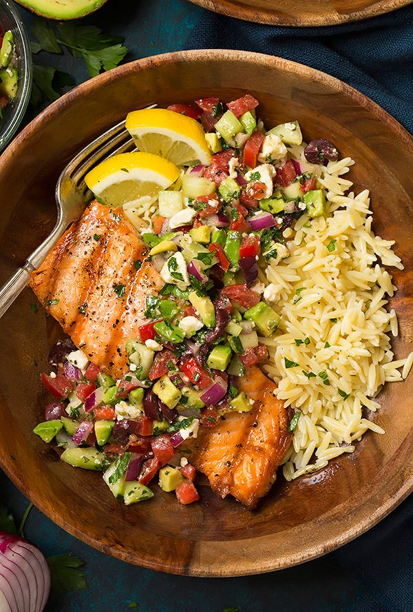 Grilled Salmon with Avocado Greek Salsa Grilled Salmon in a wooden bowl served with with Avocado Greek Salsa and a side of orzo. It's set atop a blue surface.
