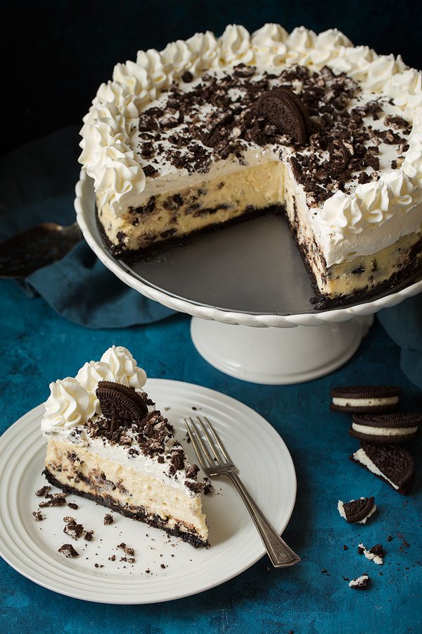 Oreo Cheesecake One slice of oreo cheesecake in low left corn of image. Whole cheesecake in upper right corner of image on a cake stand.