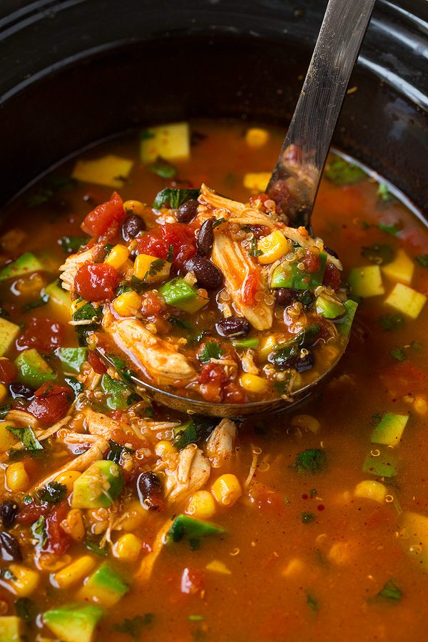 Slow Cooker Chicken Enchilada Soup Close up image of chicken enchilada soup in a ladle in center of slow cooker. Ingredients showing include shredded chicken, black beans, corn, tomatoes, avocado and spiced broth.