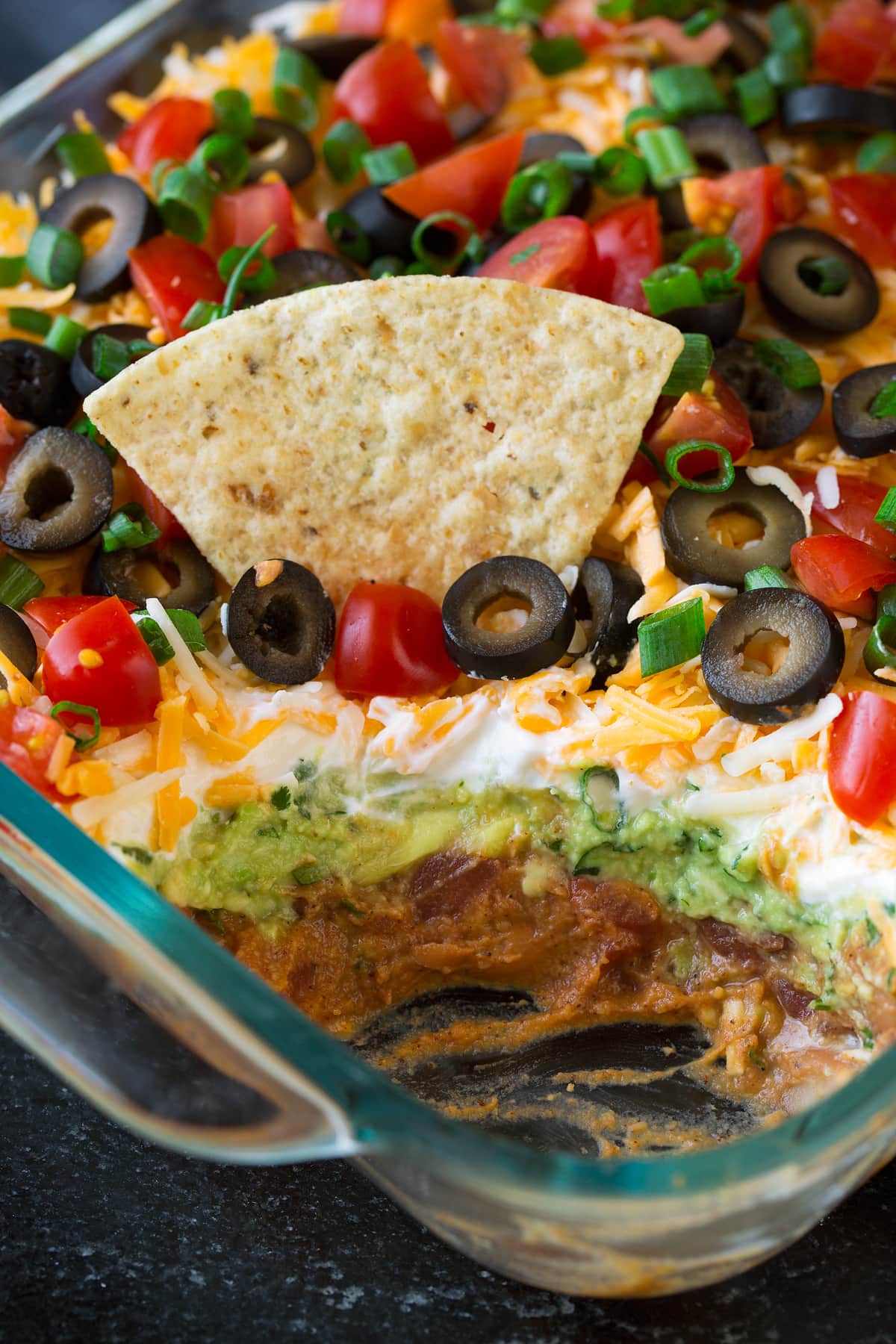 7 Layer Bean Dip - one of the all time best party foods! This stuff is addictive! #7layerdip #dip #appetizer #snack #partyfood 7 Layer Dip showing layer so of refried beans, guacamole, sour cream, cheese, olives, tomatoes and green onions.