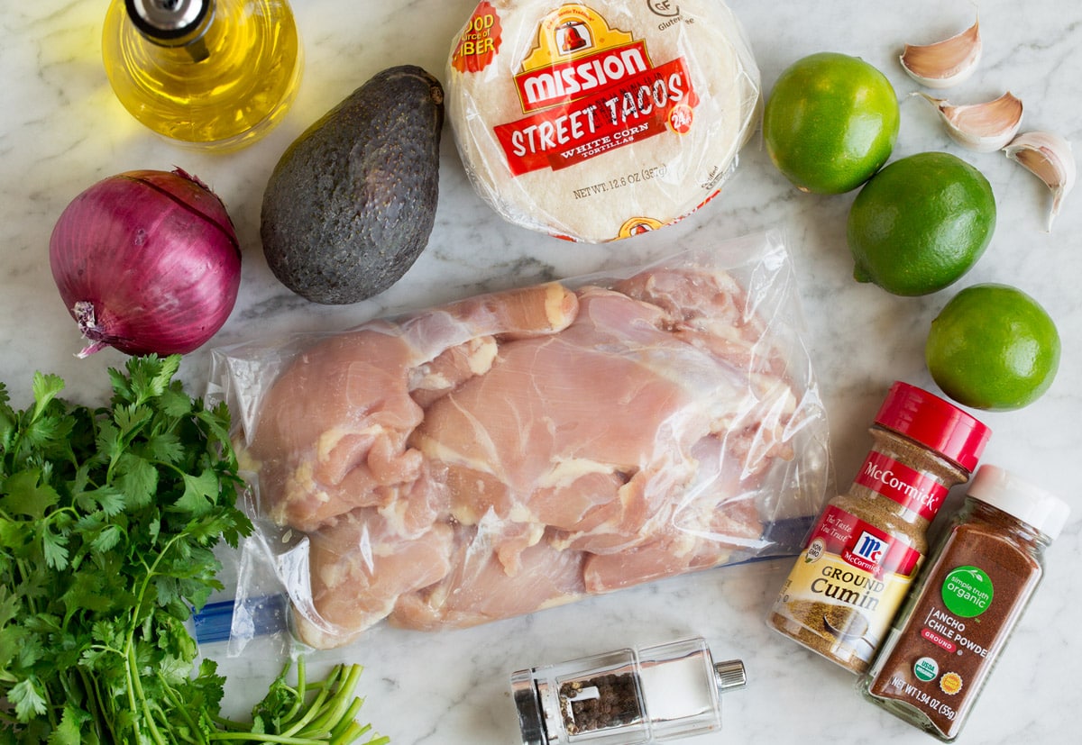 Grilled Chicken Street Tacos Image of ingredients used to make street tacos. Shows chicken thighs, cilantro, limes, garlic, avocado, chili powders, red onion, oil and tortillas.