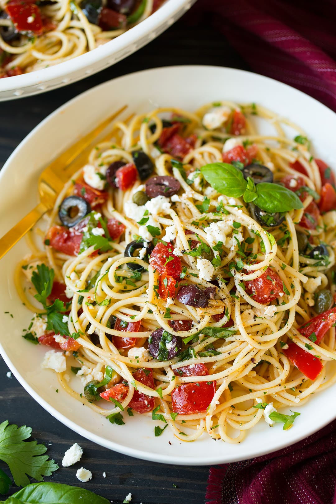 Greek Spaghetti Greek spaghetti layered with tomatoes, black olives, kalamata olives, feta, basil, garlic, olive oil. Served in a pasta bowl on a brown wooden surface.