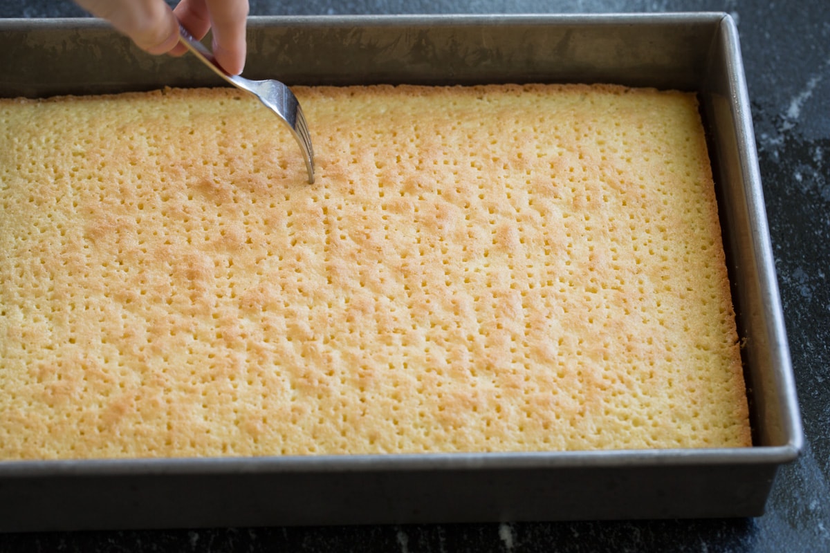 Tres Leches Cake - one of the BEST Mexican desserts! A sponge-type cake is soaked in a blend of rich milks, covered with whipped cream and dusted with cinnamon or served with strawberries. So good! #tresleches #cake #Mexicanrecipe #dessert Poking baked tres leches cake with a fork all over.