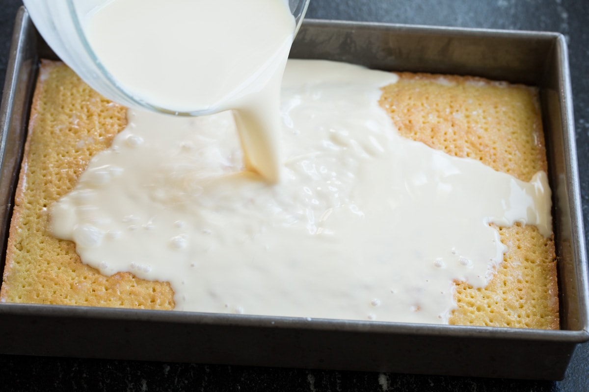 Tres Leches Cake - one of the BEST Mexican desserts! A sponge-type cake is soaked in a blend of rich milks, covered with whipped cream and dusted with cinnamon or served with strawberries. So good! #tresleches #cake #Mexicanrecipe #dessert Pouring milk mixture over poked cake.