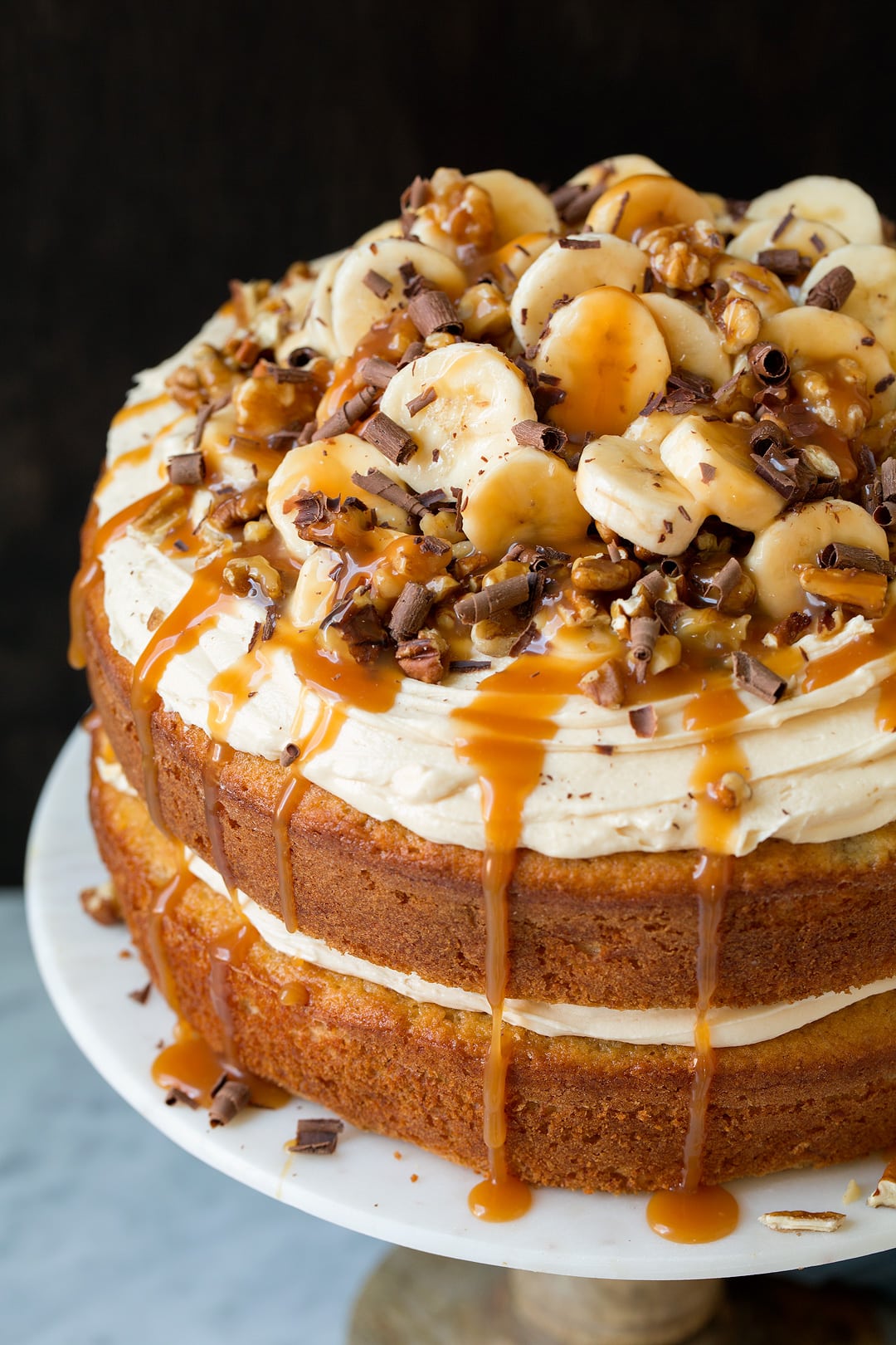Banana Cake with Cream Cheese Frosting Banana cake on a cake stand. Cake is layered with cream cheese, caramel sauce, walnuts, banana slices and chocolate shavings.