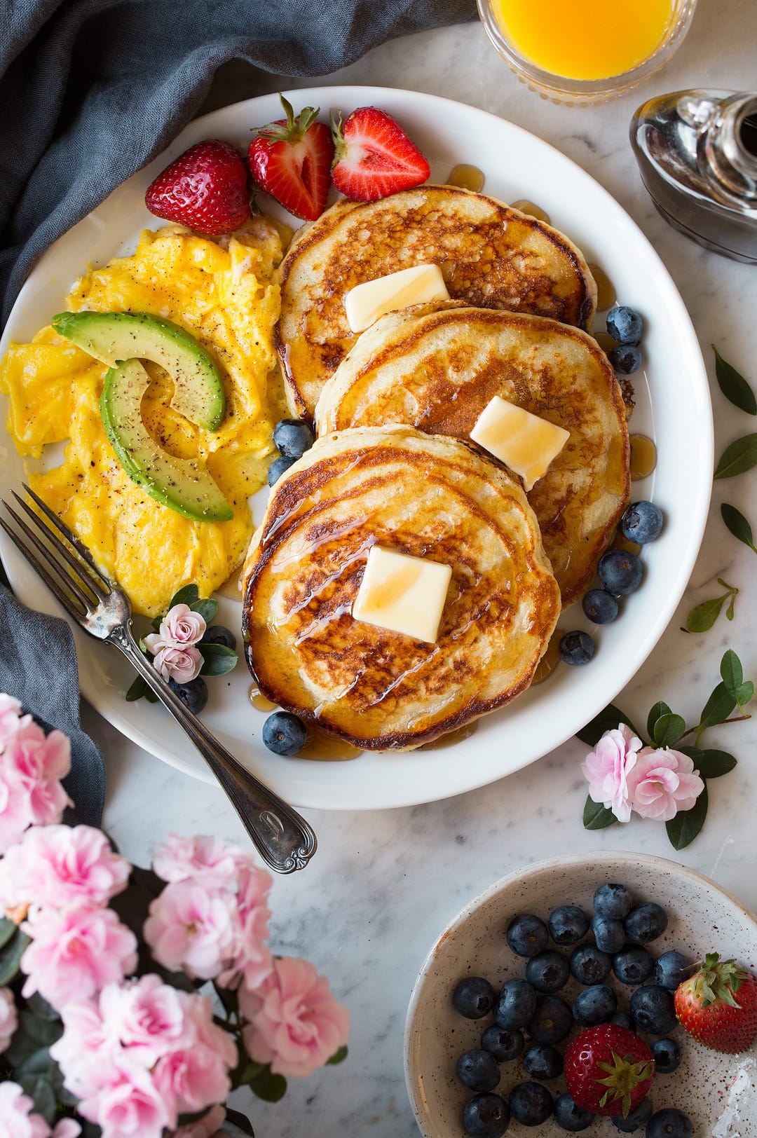 Buttermilk Pancakes Three buttermilk pancakes on a large white plate dotted with slabs of butter and drizzled with maple syrup. Included on the side are scrambled eggs and fresh fruit.