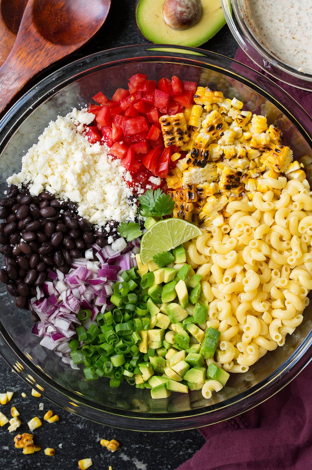Mexican Macaroni Salad Mexican macaroni salad ingredients in a large glass bowl before tossing. Ingredients include macaroni, charred corn, black beans, avocado, tomato, green onion, red onion, and cotija.