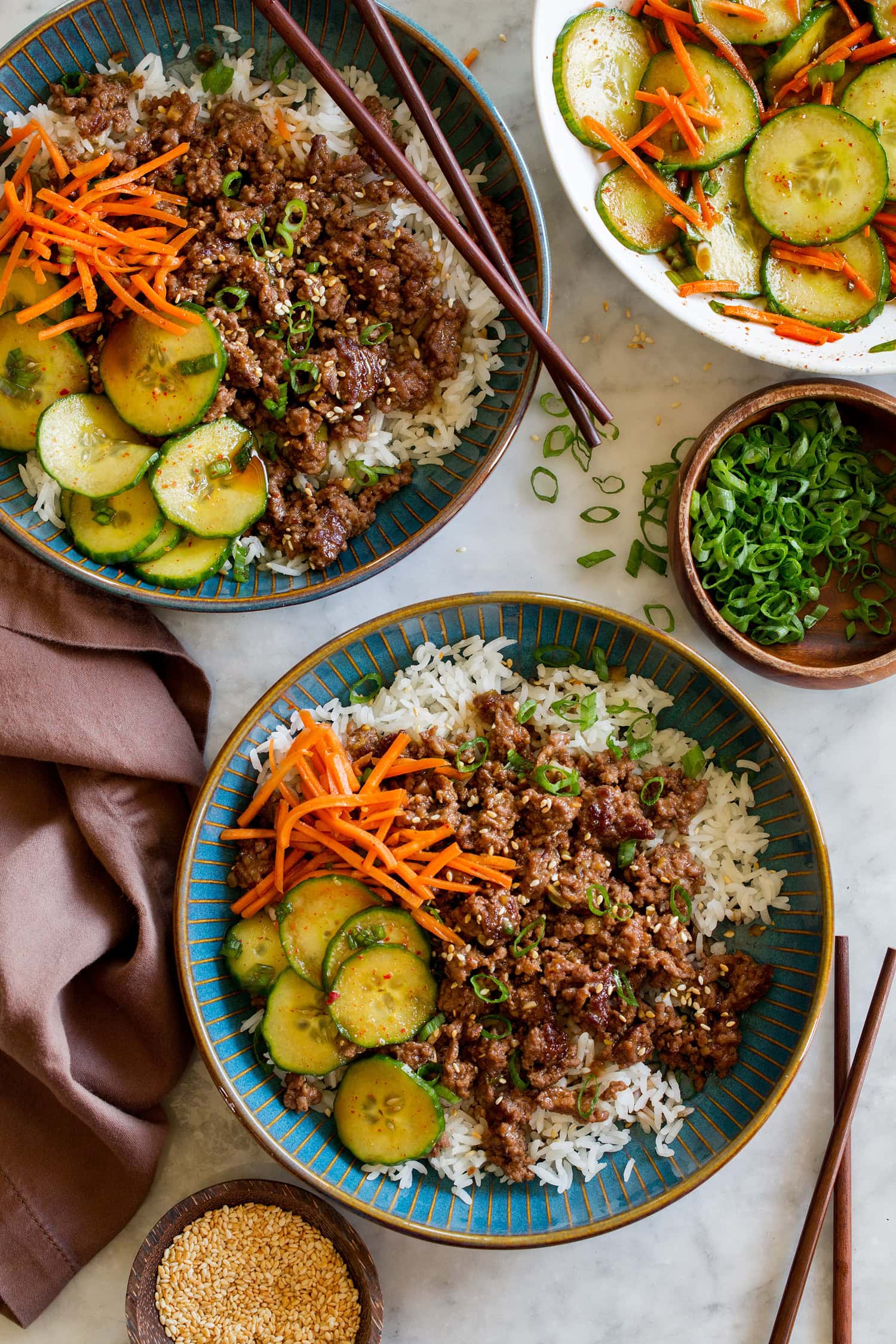 Korean Beef Bowls Korean beef bowls shown with serving suggestion of sides of Korean vegetables including cucumbers and carrots.
