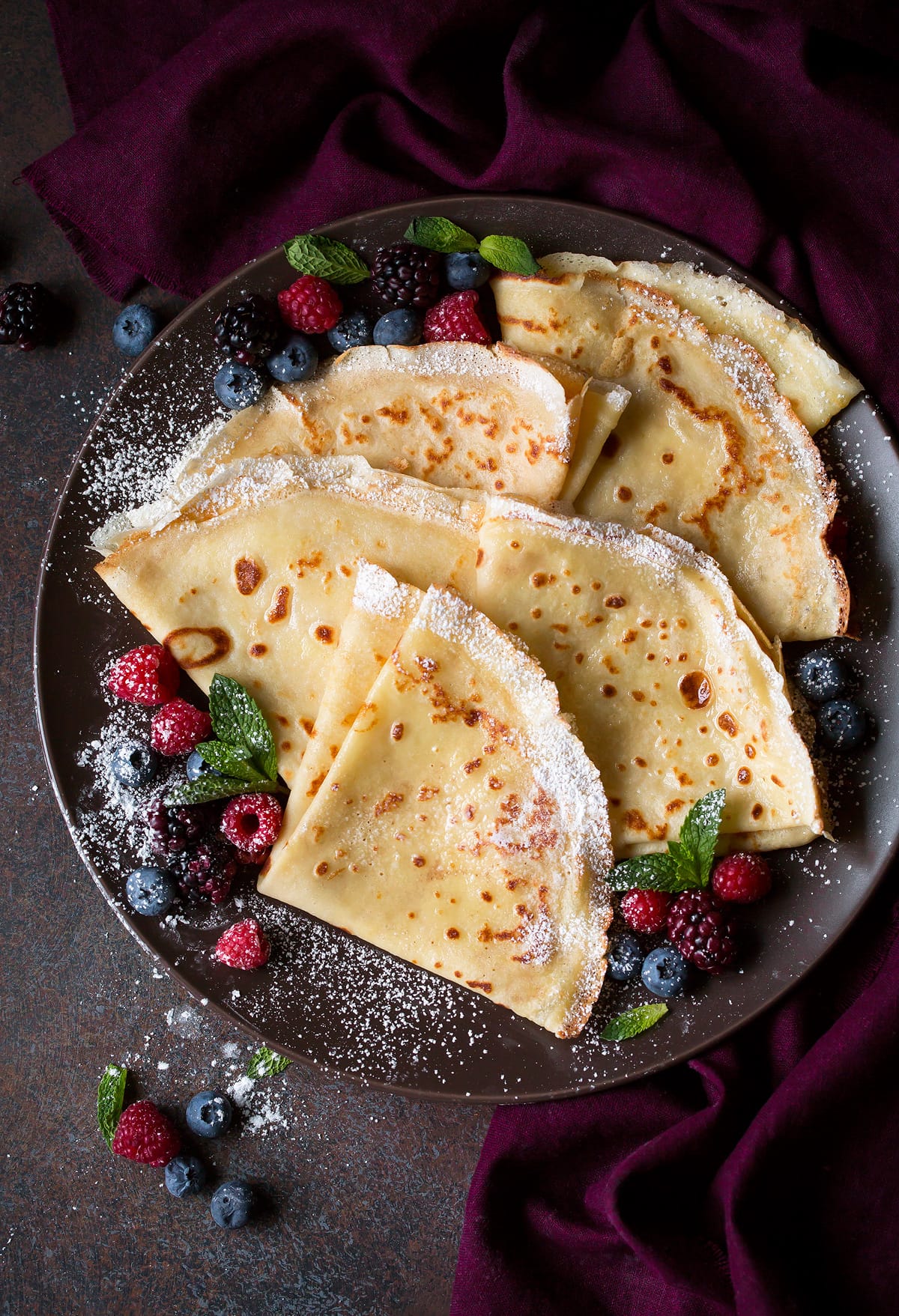 Crepes Recipe Overhead image of plate full of folded crepes on a dark plate set over a maroon napkin.