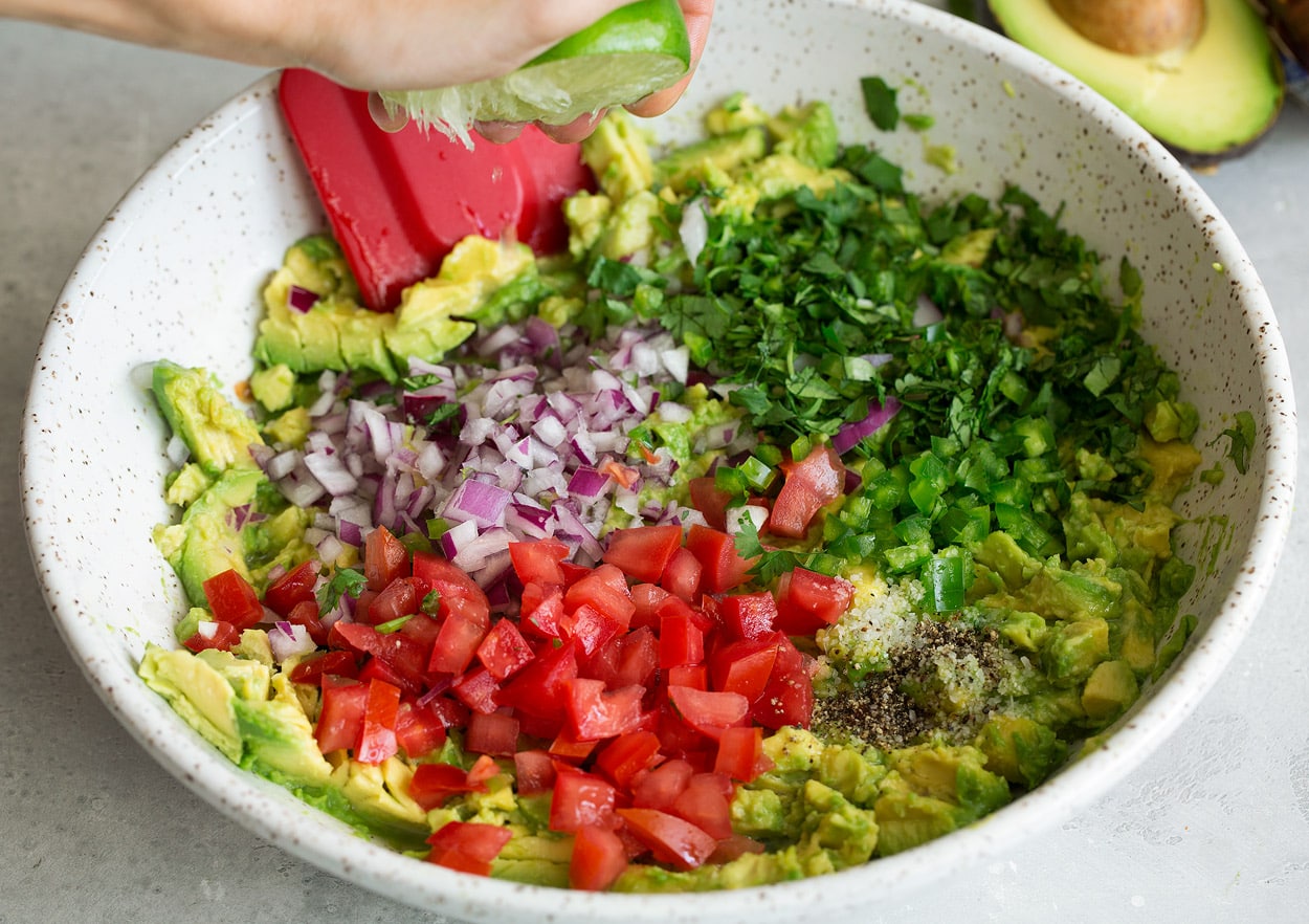 Homemade Guacamole Adding diced tomatoes, red onion, cilantro, garlic, jalapeño and lime juice to mashed avocado in mixing bowl.