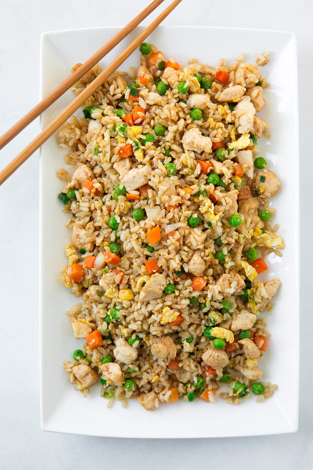 An easy flavorful fried rice that's made with brown rice and lean chicken breast instead of white rice and ham. A recipe everyone can agree on! Hearty and so satisfying. It make great leftovers too. | Chicken Fried Rice | Chinese Take Out | #cookingclassy #rice #chicken An overhead image of chicken fried rice on a white platter with chop sticks on the side.
