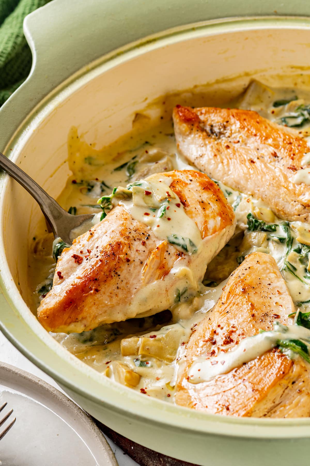 Creamy spinach artichoke chicken is a fast and easy one-pan dinner recipe brimming with briny artichokes, spinach, and lots of melty cheese. A spoon lifting a saucy chicken piece from creamy spinach artichoke chicken in a large skillet.
