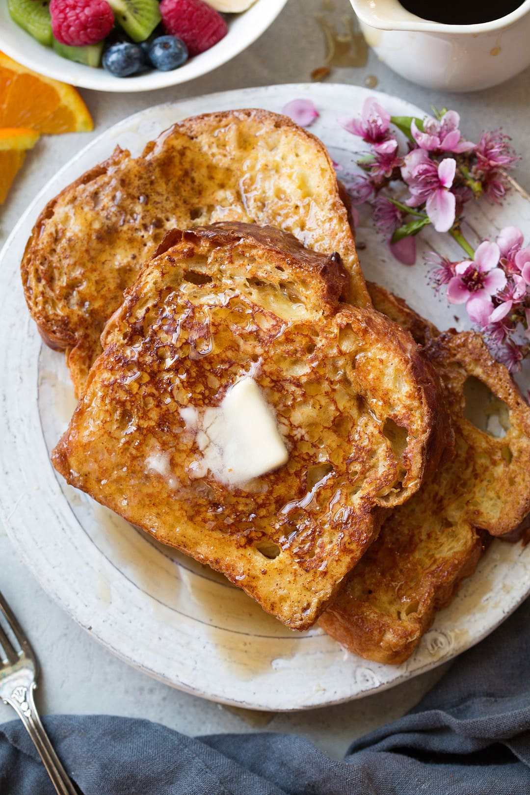 The BEST French Toast! Three slices of french toast laying together on a white plate. Topped with butter and maple syrup and garnished with flowers on the side and fresh fruit.