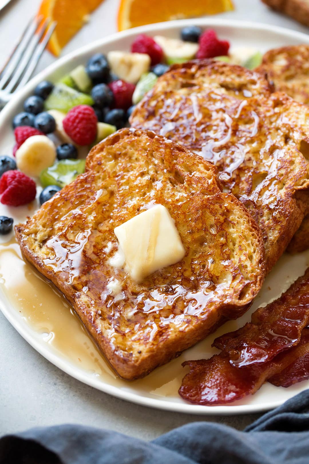 The BEST French Toast! Row of french toast on a plate topped with maple syrup and butter and served with a side of bacon.