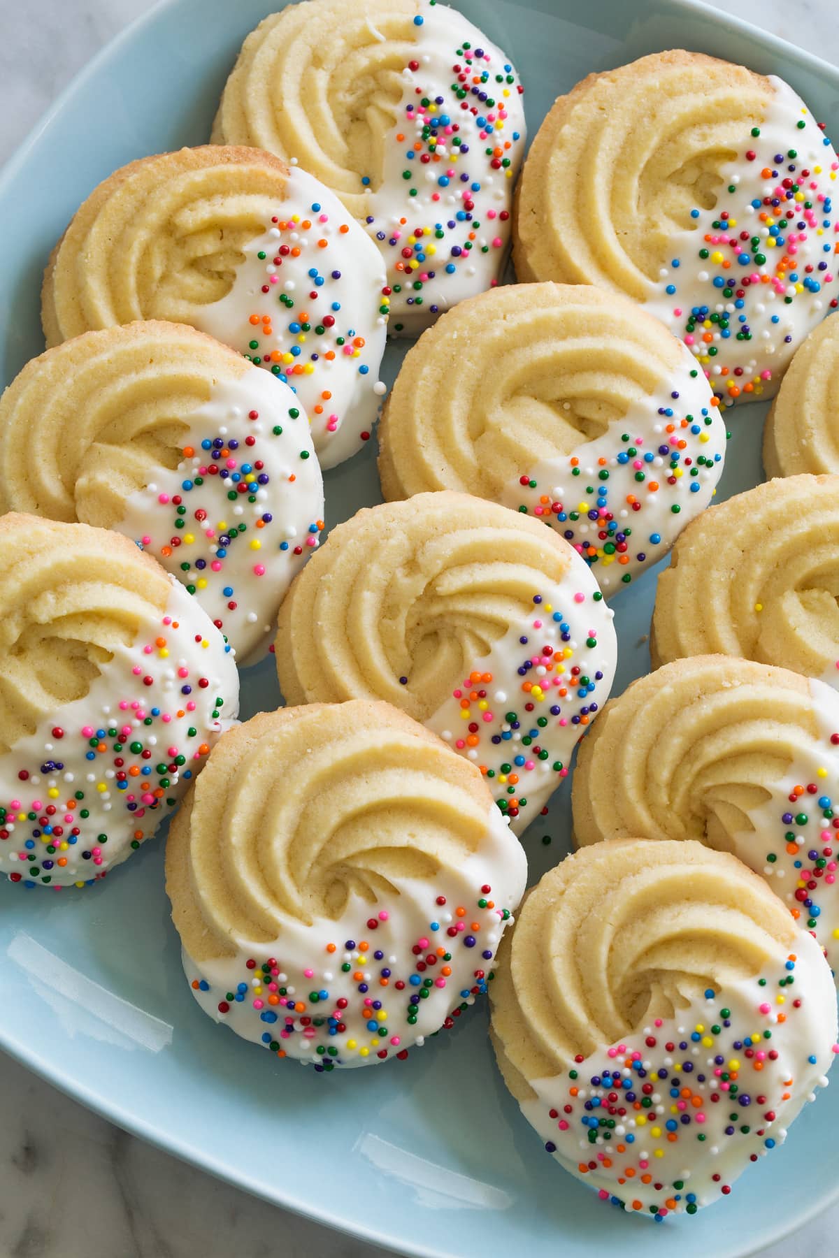 Butter Cookies Butter Cookies dipped in white chocolate and covered with rainbow sprinkles. Set in three rows on a turquoise plate.
