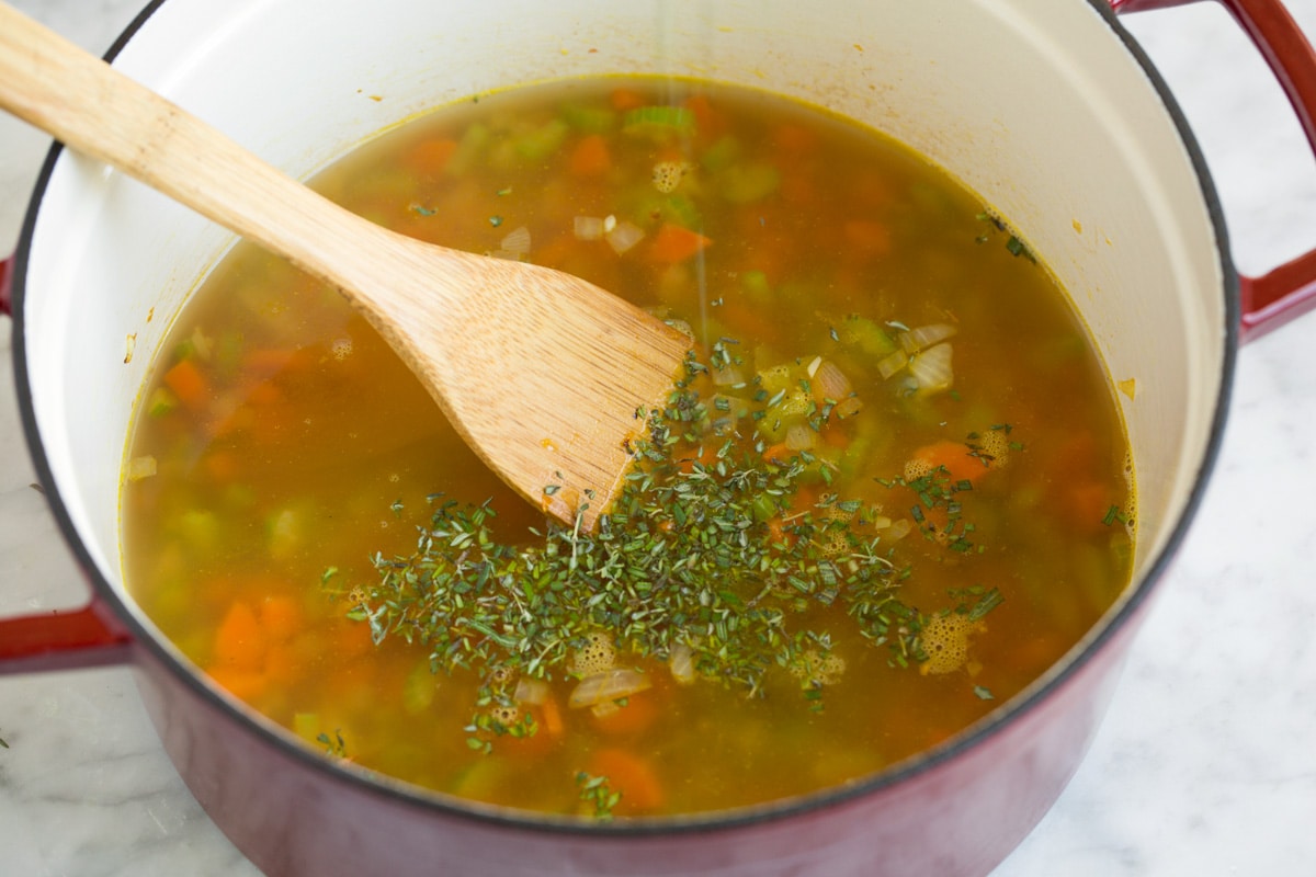 Creamy Chicken Gnocchi Soup Adding broth and fresh herbs to soup mixture in pot.