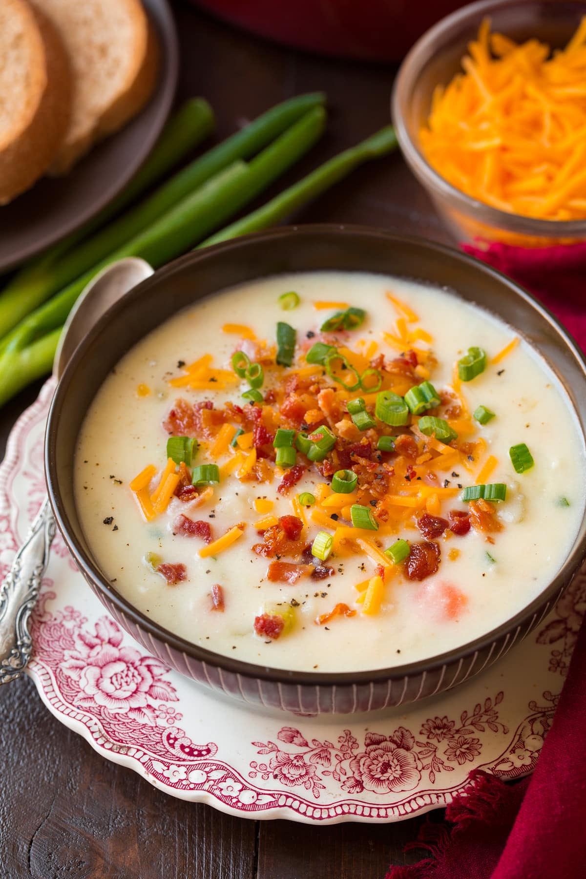 Potato Soup Photo: Serving of potato soup shown in a brown bowl set over a decorated white and red plate. Potato soup is garnished with cheddar, bacon and green onions. Wheat bread is shown as a serving suggestion.