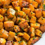 Roasted butternut squash with garlic and fresh herbs in a white ceramic serving bowl set over a white marble surface.