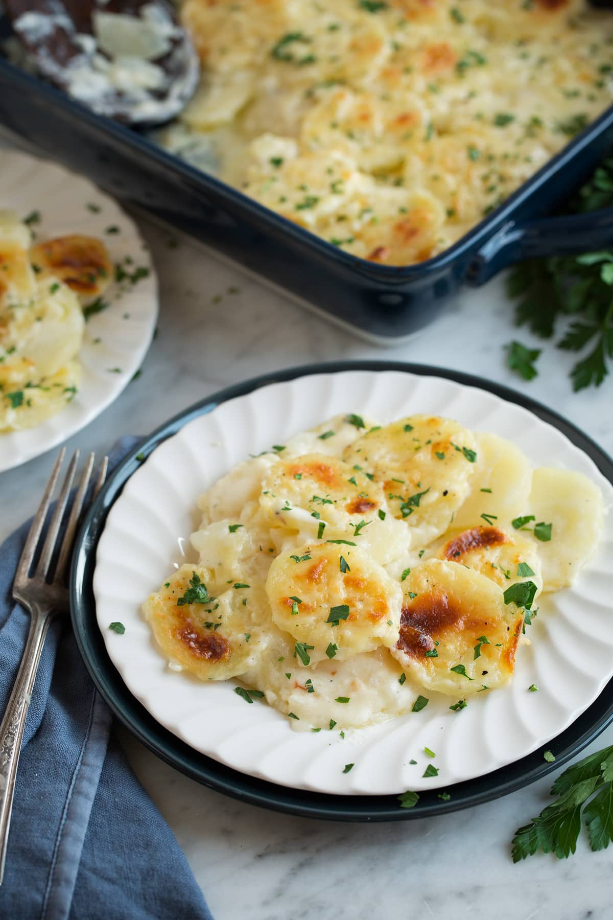 Scalloped Potatoes Scalloped Potatoes on a white single serving dish set over a dark blue plate that's resting on a marble surface.