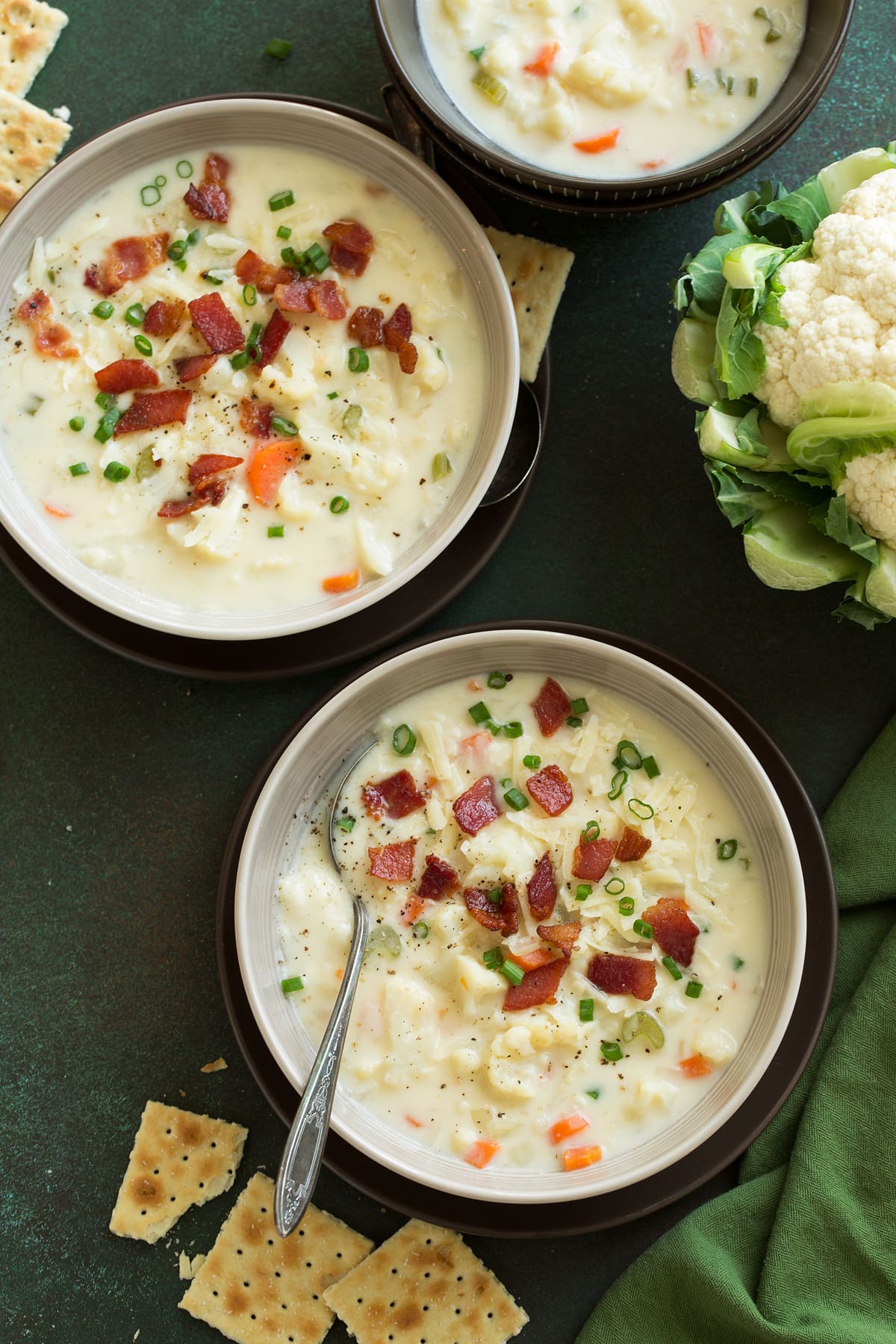 Cauliflower Soup Three servings of Cauliflower Soup in grey bowls set over brown plates on a green tabletop surface.