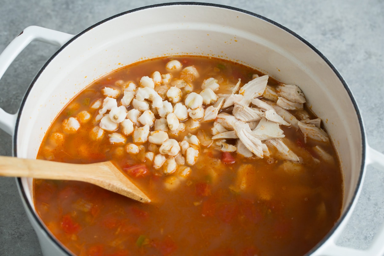 Showing how to make chicken posole. Adding chicken and hominy to pot of soup.