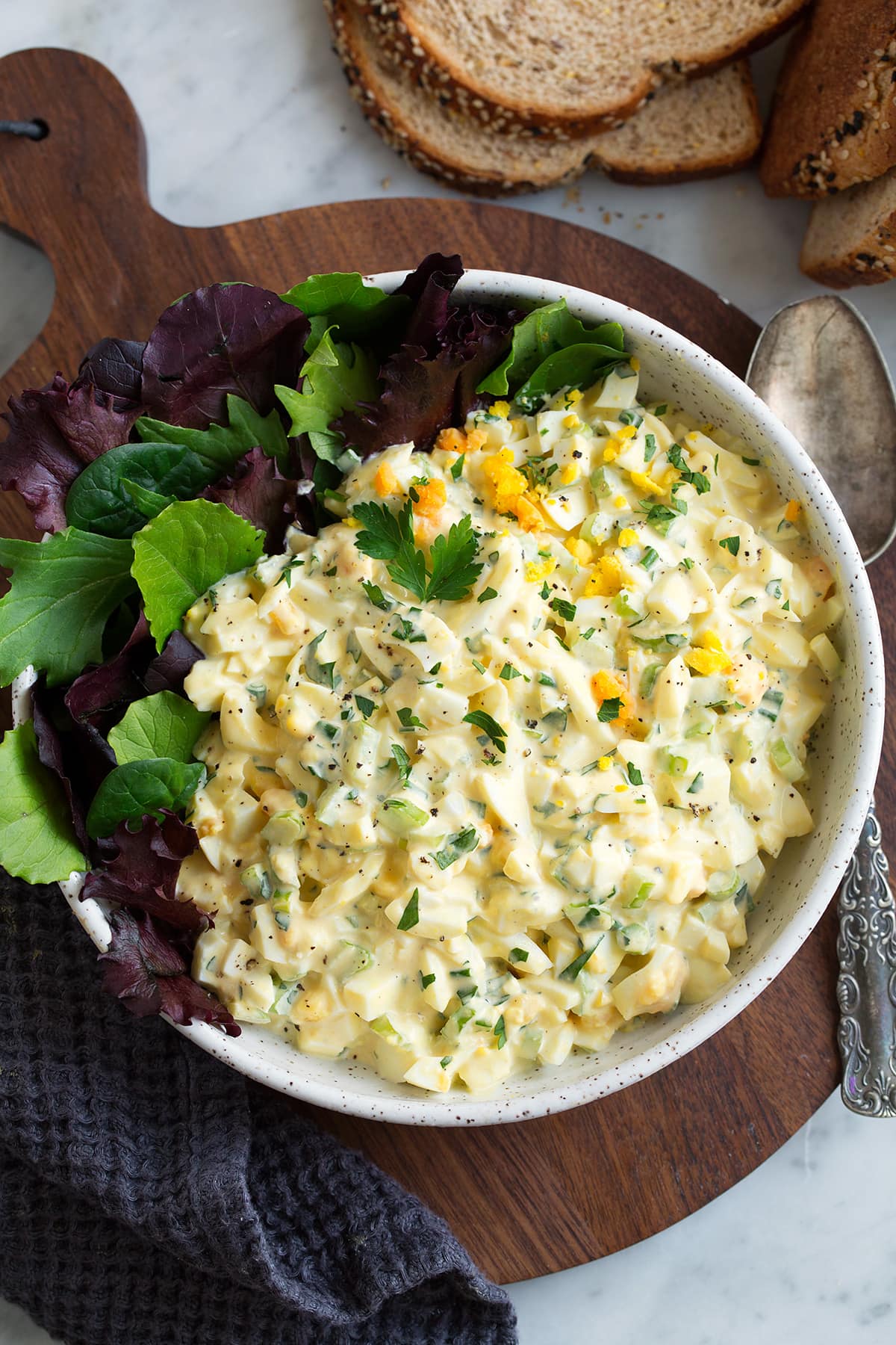 Egg Salad Egg Salad in a white speckled ceramic serving bowl with a side of colorful lettuce. Bowl is sitting on a wooden platter which is on a marble tabletop. There is a silver spoon of the the side and whole grain bread in the corner.