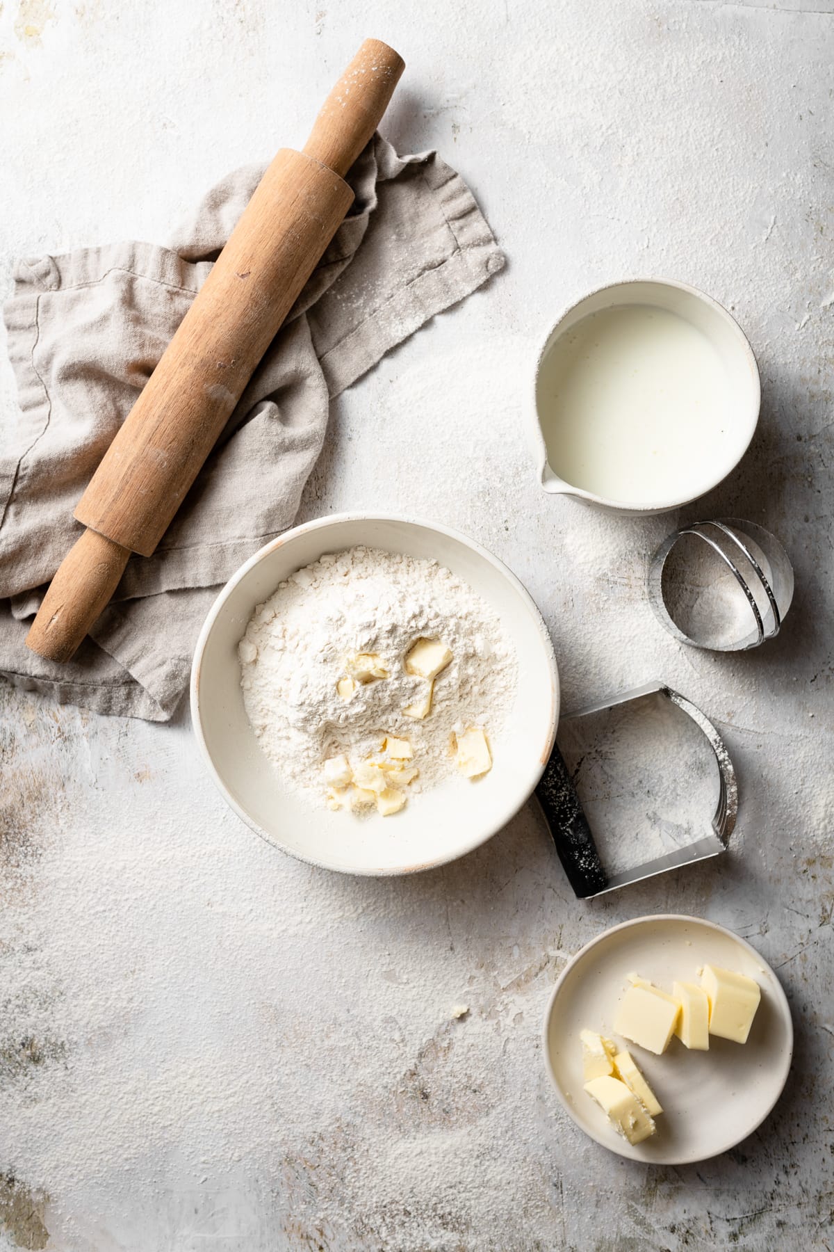 Buttermilk Biscuits Showing biscuit ingredients including flour mixture, butter and buttermilk in mixing bowls. Rolling pin on the side and beige kitchen towel.