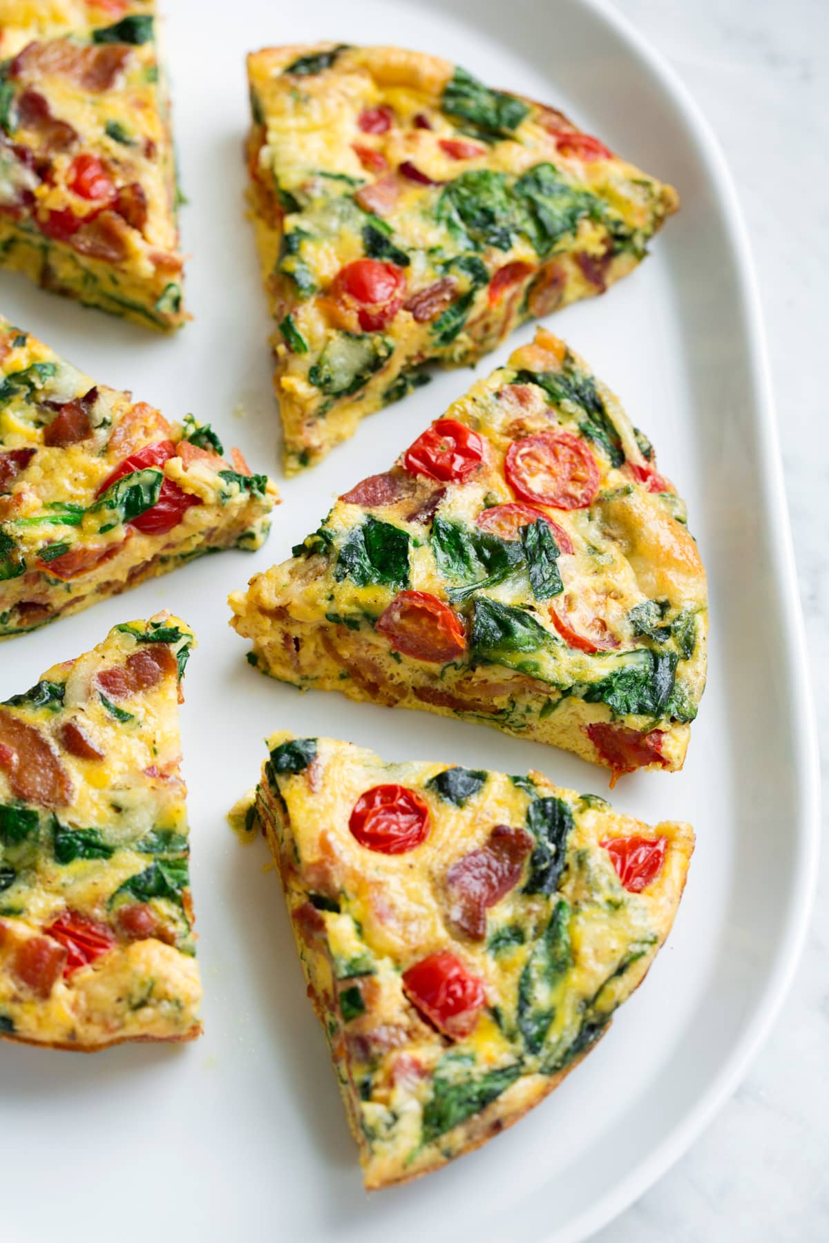 Frittata Recipe (Bacon, Spinach, Tomato and Swiss) Six frittata wedges shown on a white serving platter set over a marble surface. Frittata is filled with bacon, spinach, tomatoes and swiss.
