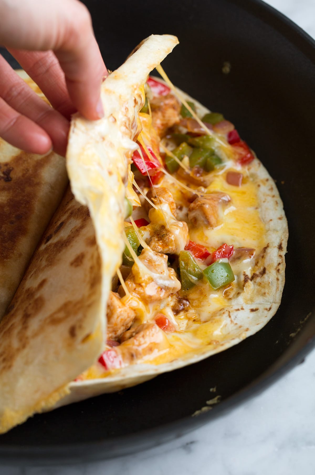 Loaded Chicken Quesadillas Hand opening quesadilla in a skillet to show filling of cheese, bell peppers, onions, and seasoned chicken pieces.