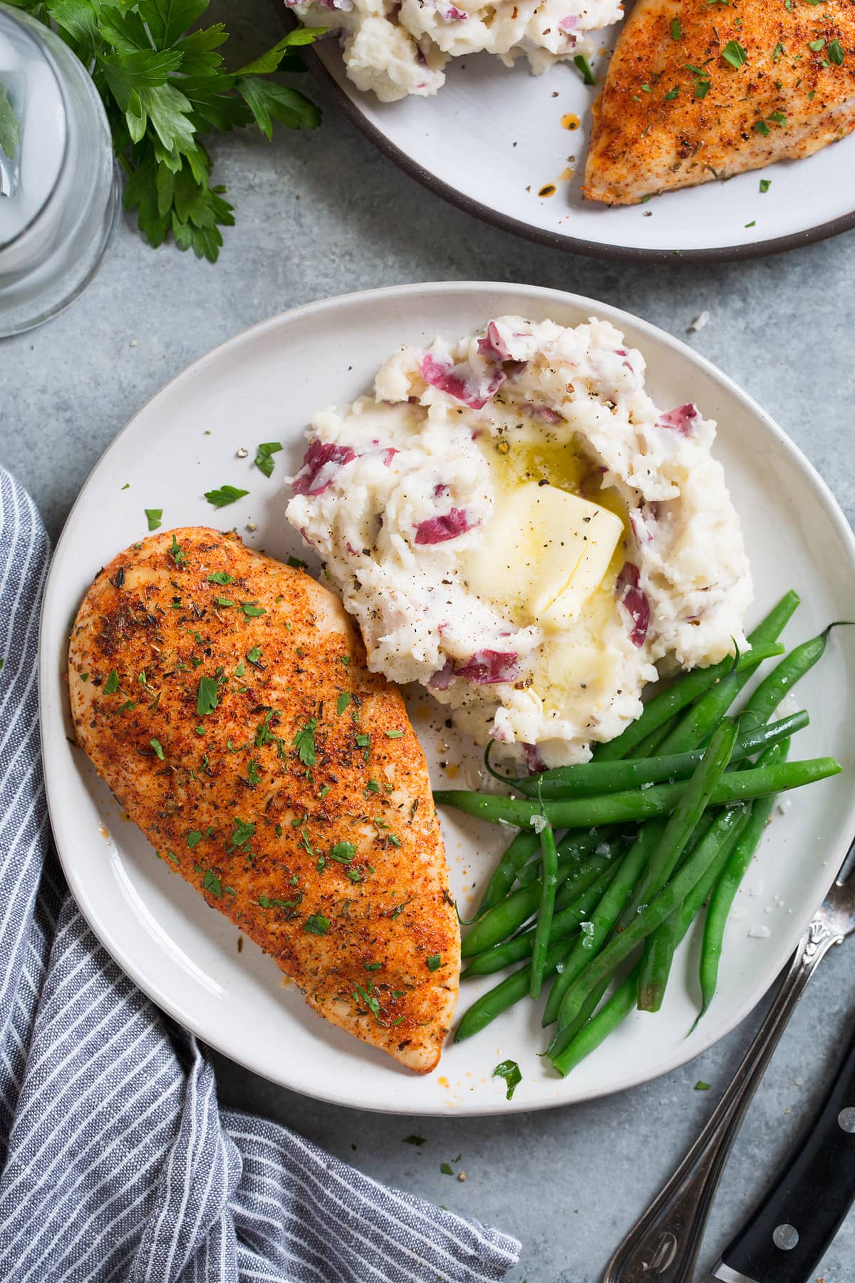 Baked Chicken Breasts Overhead image of a seasoned baked chicken breast on a white serving plate along with a side of mashed potatoes and steamed green beans.
