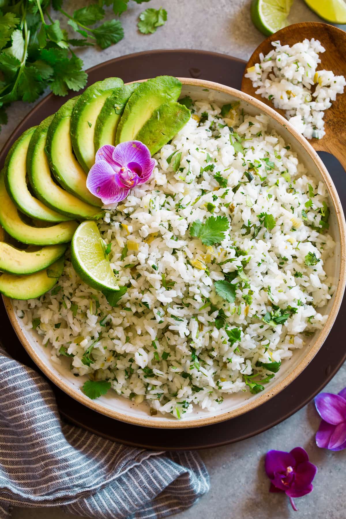 Cilantro Lime Rice Cilantro Lime Rice in a cream ceramic serving bowl set over a brown matte plate. Rice is garnished with cilantro, lime, avocado and orchid flower.