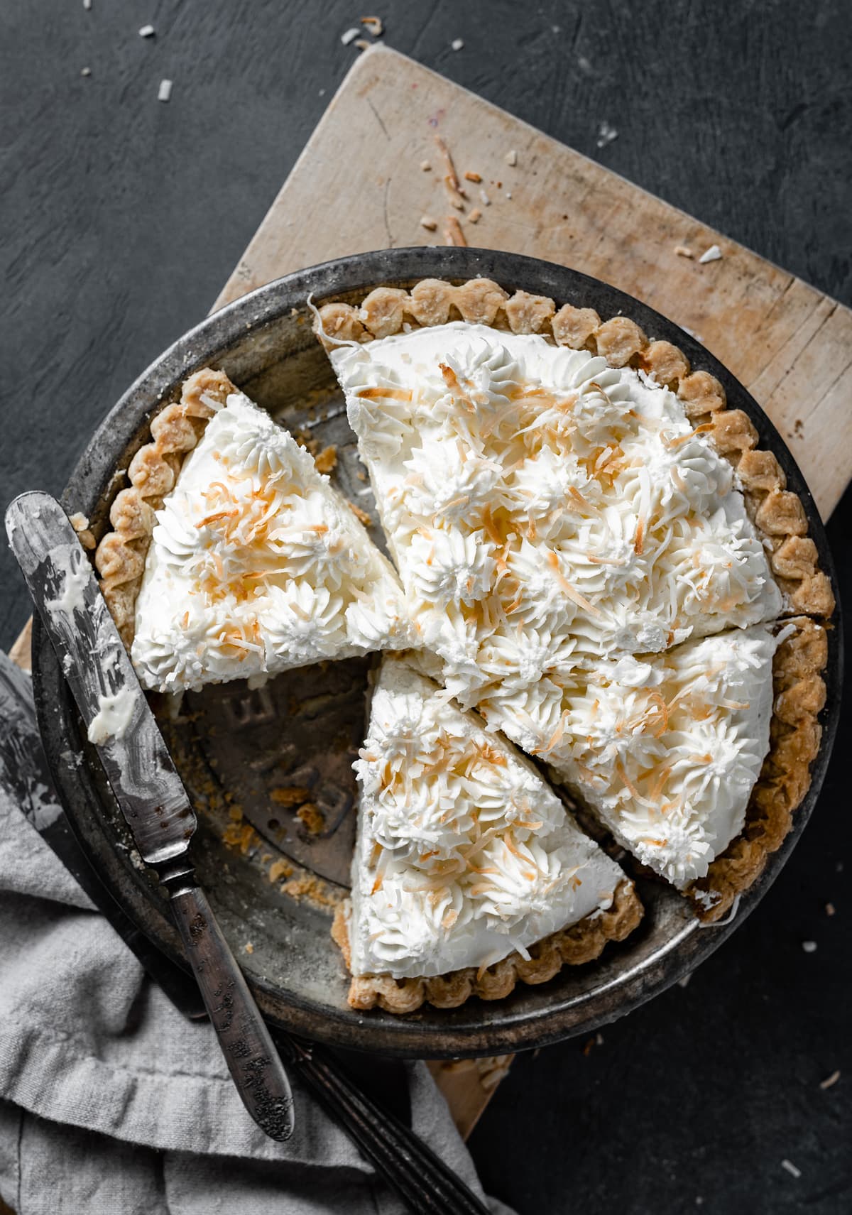 Coconut Cream Pie Whole coconut cream pie in a pie dish with slices cut. Pie is sitting on a wooden cutting board over a dark surface.