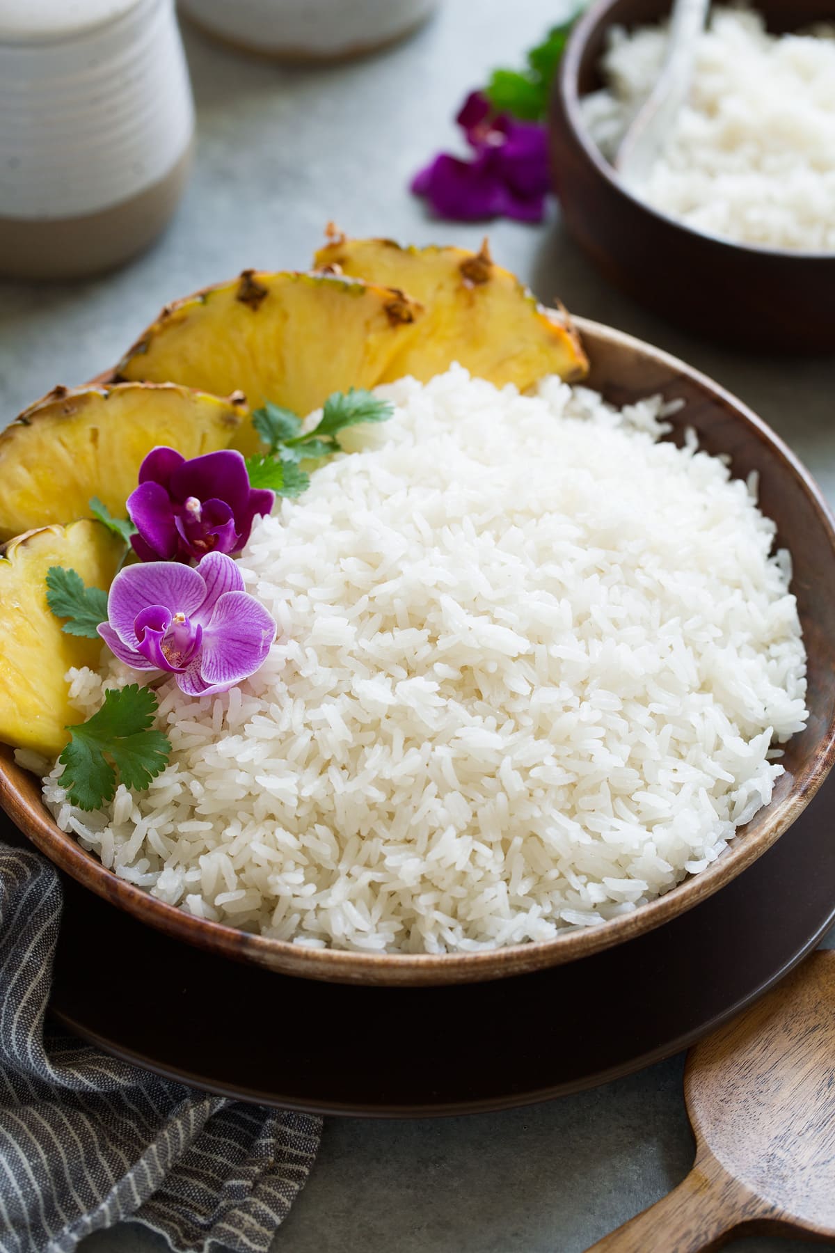 Coconut Rice Recipe Coconut rice in a wooden serving bowl garnished with orchids, cilantro and pineapple wedges on the side.