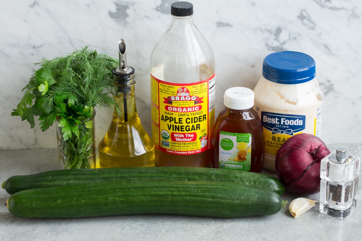 Refreshing Cucumber Salad Ingredients needed to make cucumber salad shown here including English cucumbers, mayonnaise, olive oil, garlic, apple cider vinegar, dill, parsley, honey, and red onion.