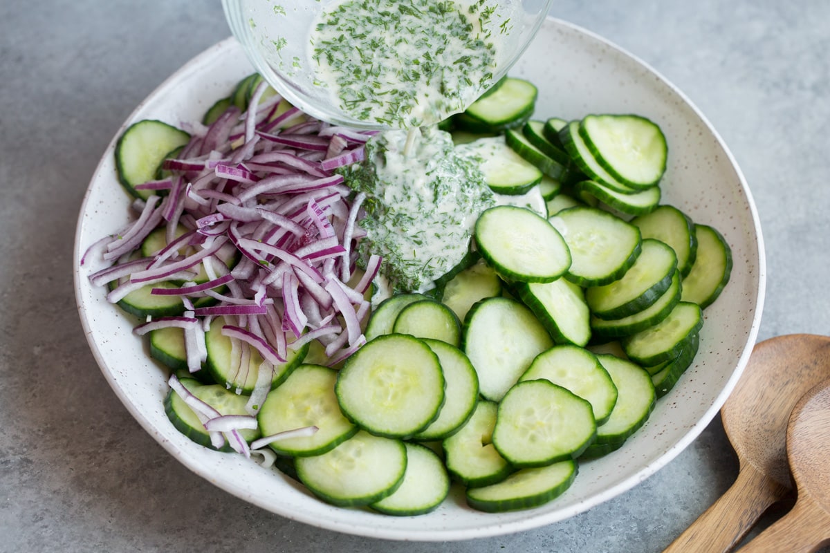Refreshing Cucumber Salad Pouring dressing over cucumbers and red onion in white serving bowl.