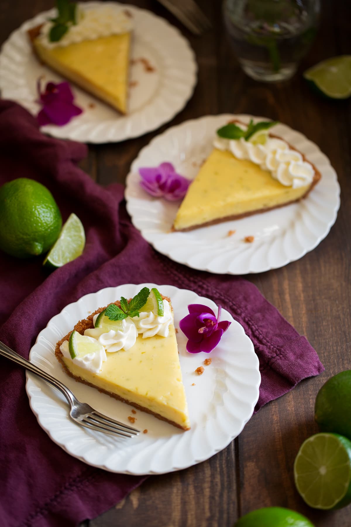 Best Ever Key Lime Pie! Three slices of key lime pie on white scalloped dessert plates set over a maroon linen napkin. Plates are decorated with purple orchids.