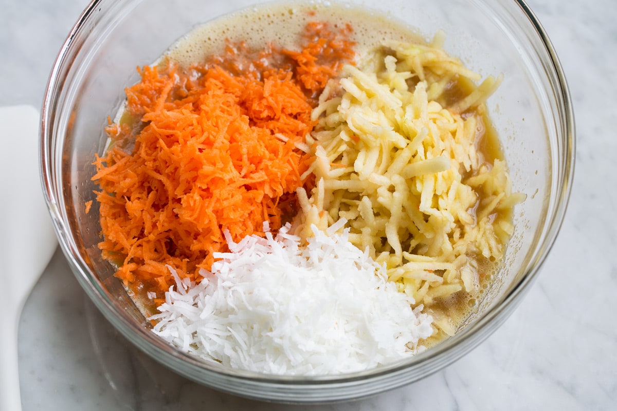 Morning Glory Muffins Adding shredded carrots, apples and coconut to morning glory muffin batter in glass mixing bowl.