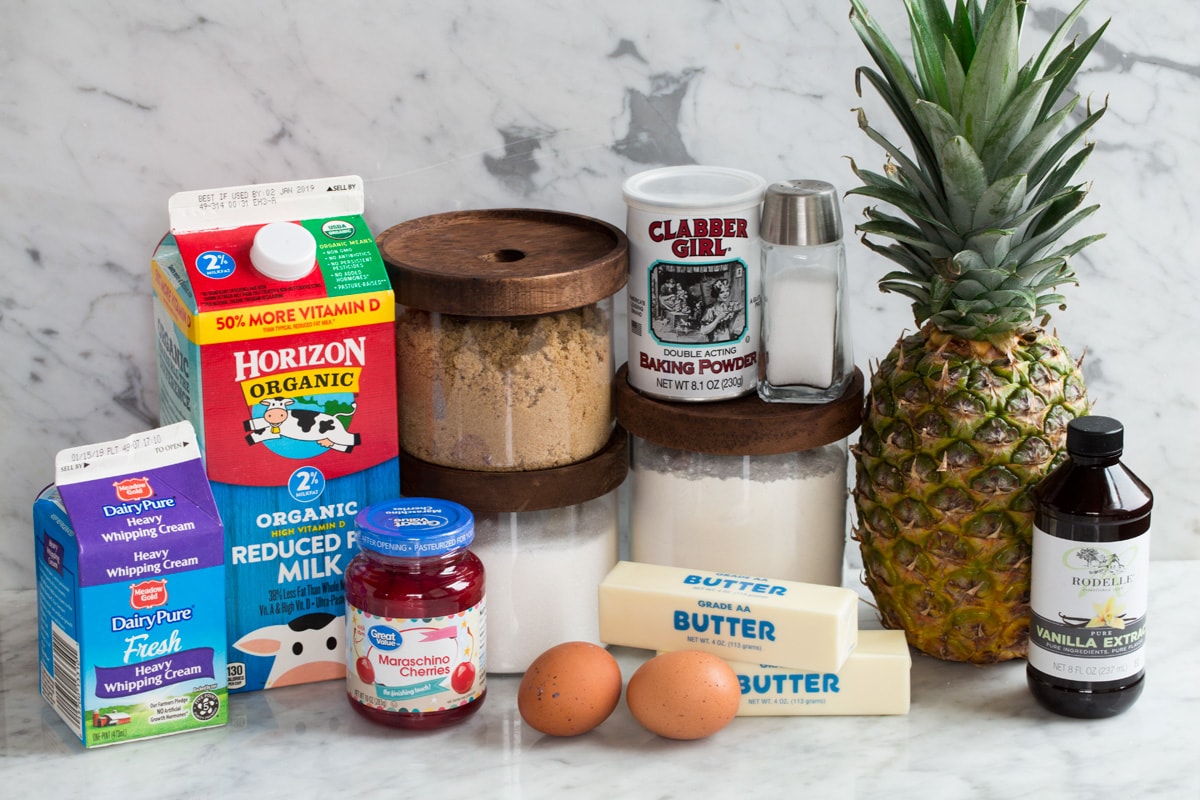 Pineapple Upside Down Cake Ingredients needed to make pineapple upside down cake shown here including fresh pineapple, flour, brown and white sugar, butter, eggs, baking powder, salt, milk, cream and maraschino cherries.