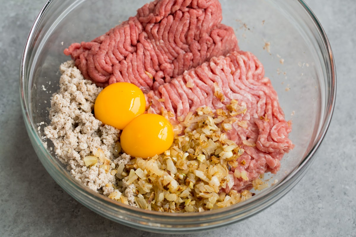 BEST Swedish Meatballs Recipe (Oven Baked)! Mixing ground beef, ground pork, bread crumb mixture, sautéed onions and egg yolks in a glass mixing bowl.